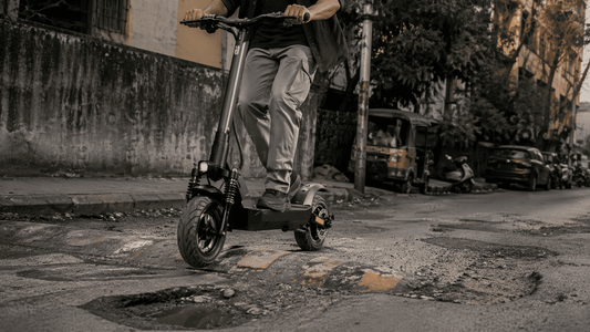 Rider on a black electric scooter crossing potholes and a speed breaker on a rough Indian street.